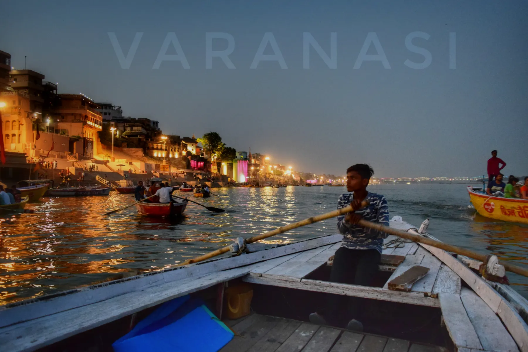 A boatman rowing past the lit ghats at dusk